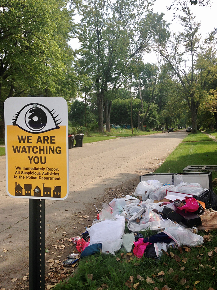 Resident-made sign in Brightmoor stating that residents are watching their block, next to illegally dumped trash on side of road. 