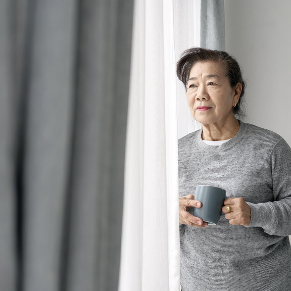 Older woman looking out window while holding mug.
