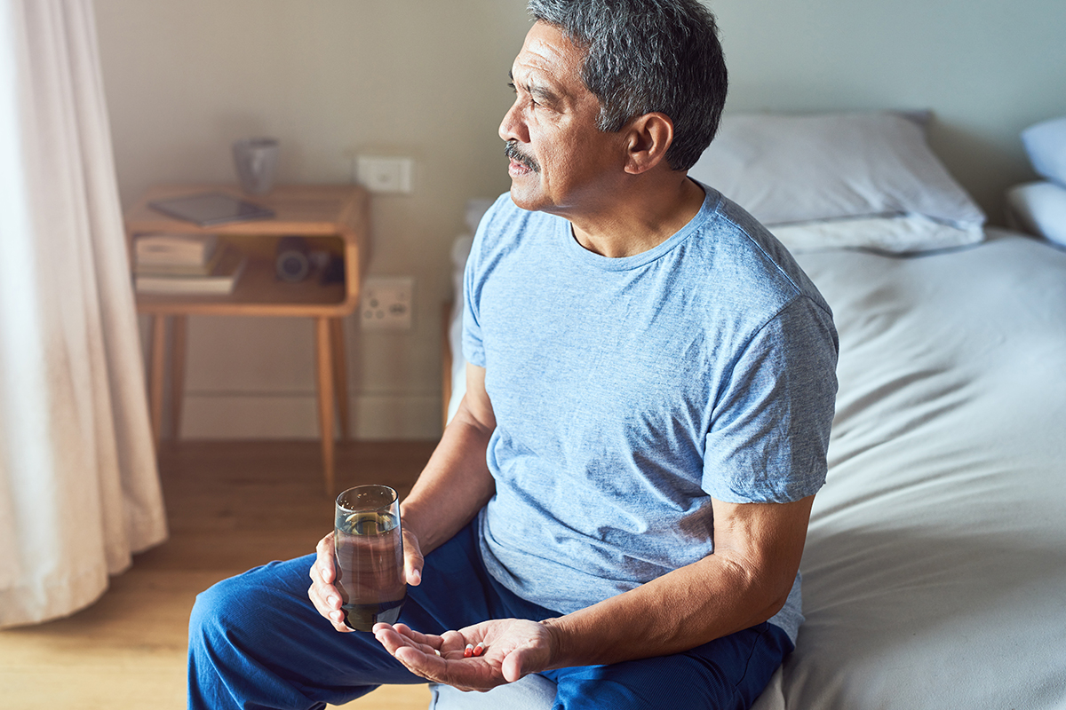 Older man sitting in bedroom with medication and glass of water.