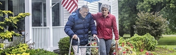 Older couple walking in front of a house, one with a walker